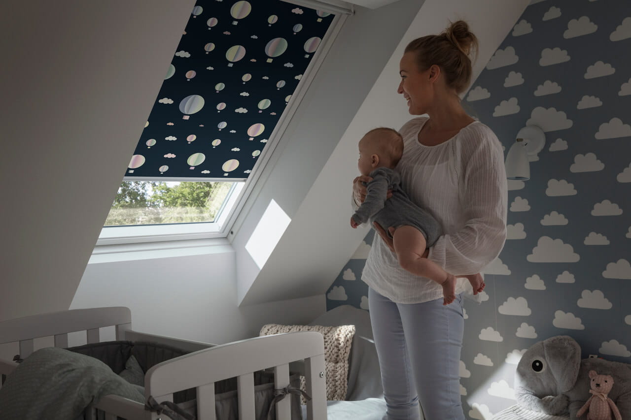 Image of a person holding a baby and looking at VELUX Disney blackout blinds installed in a window.