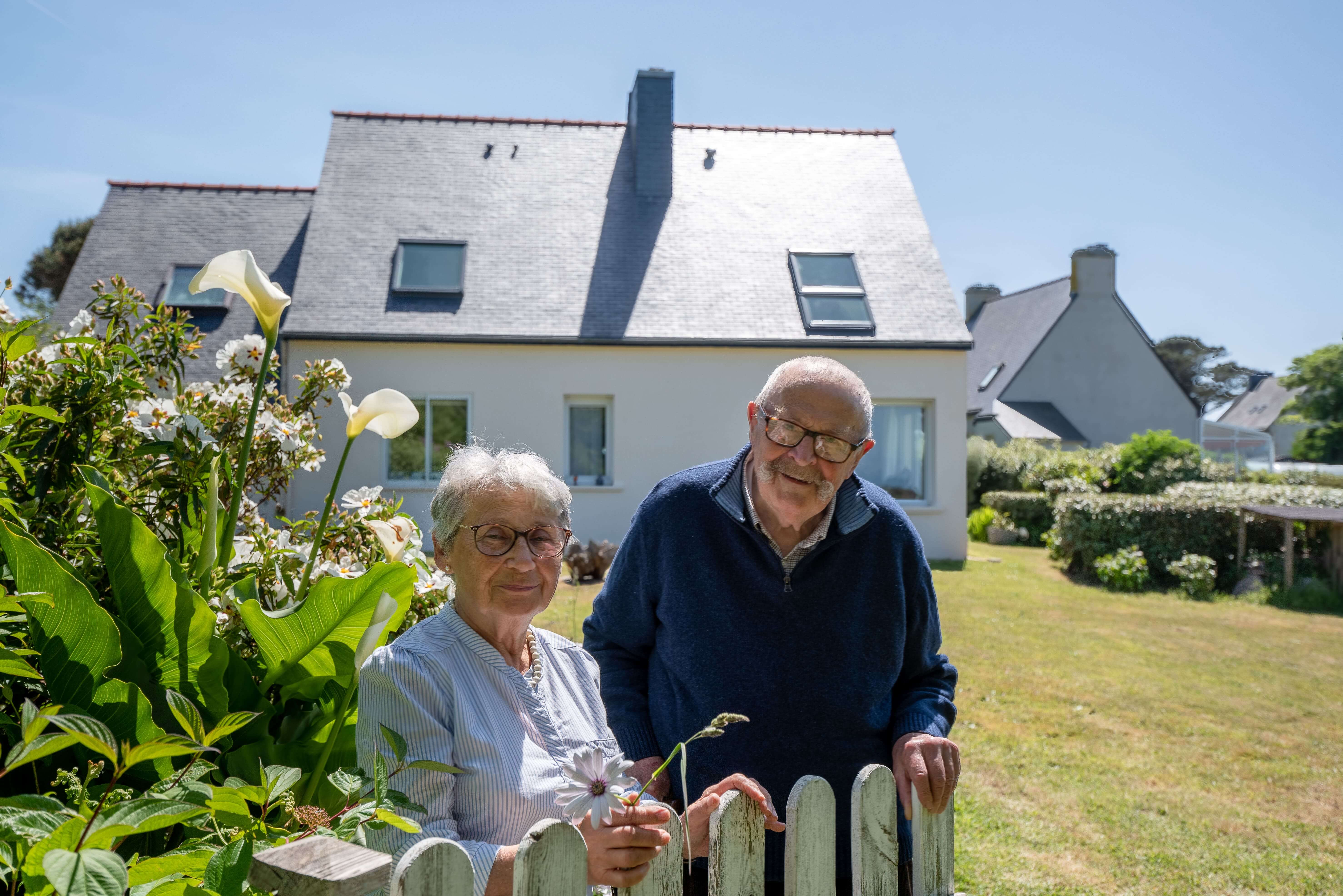 An elderly couple standing by the fence and their house is behind them